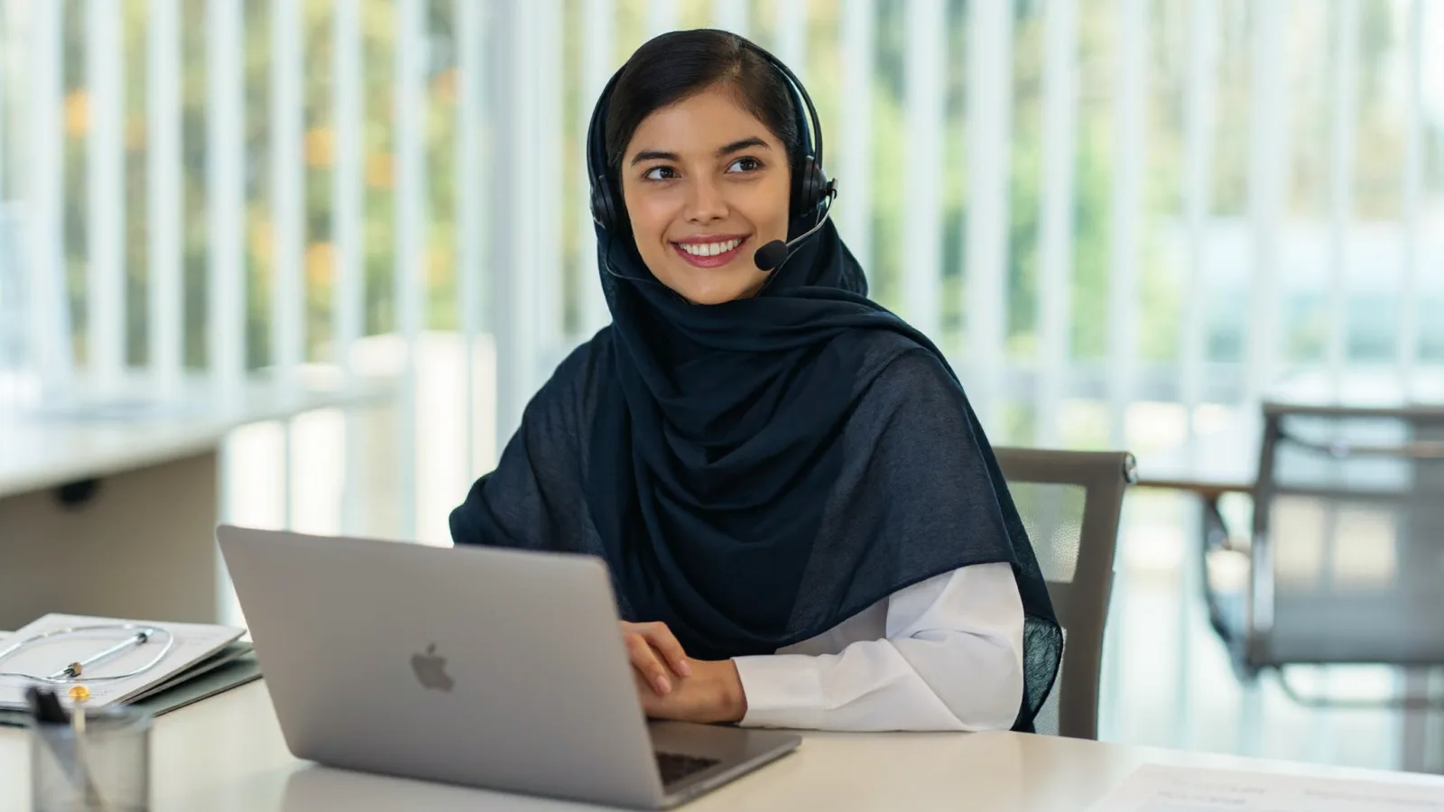 A warm, smiling Iraqi support specialist wearing a headset at a modern desk, ready to help in Arabic and English