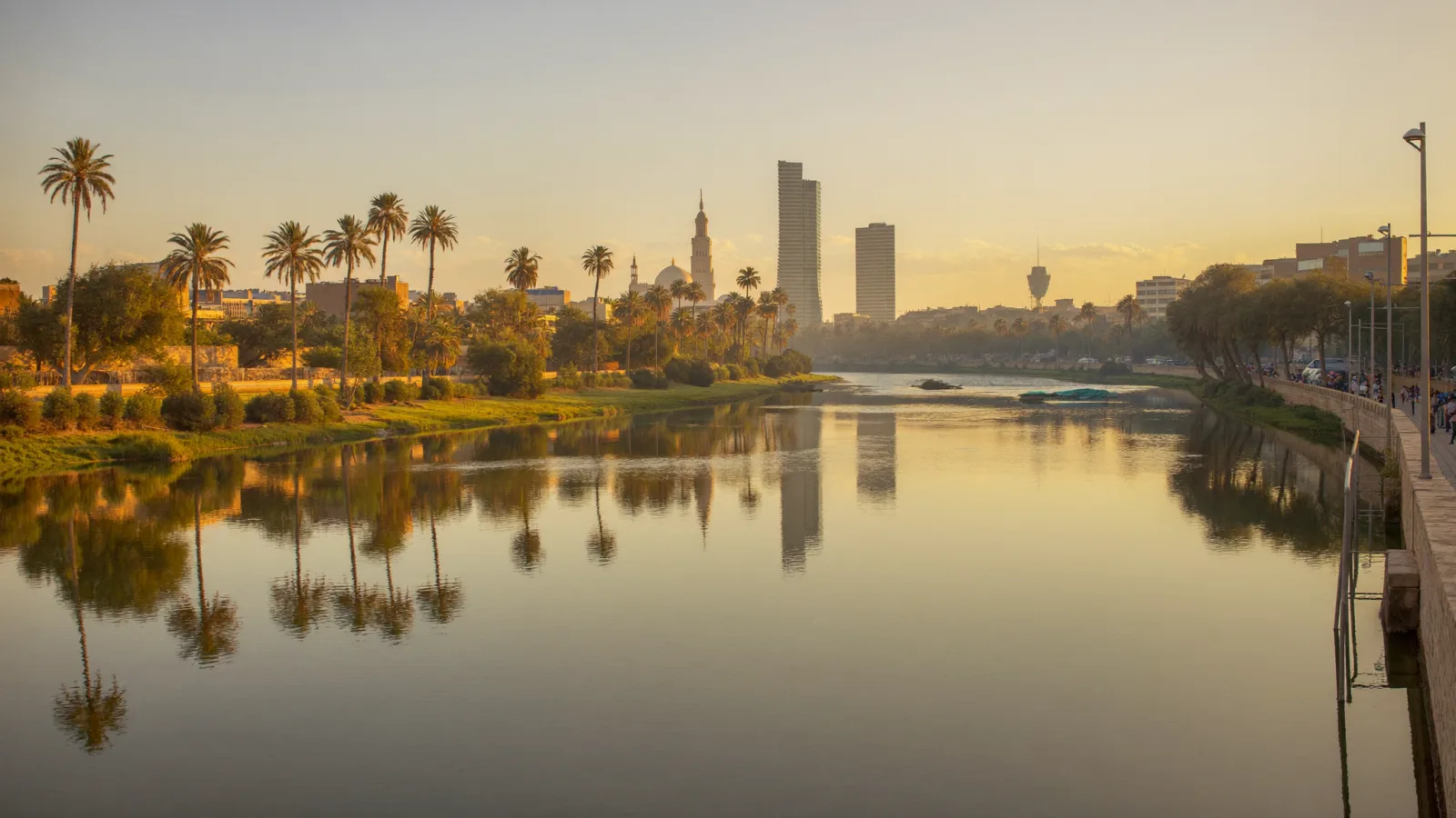نهر دجلة يجري عبر بغداد عند الغروب الذهبي، ضفافٌ تحفّها أشجار النخيل وأفق المدينة منعكسٌ على صفحة الماء الساكنة